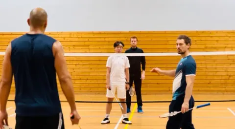 Un coach explique l’exercice à trois stagiaires adultes réunis au filet, tribunes en bois en arrière-plan