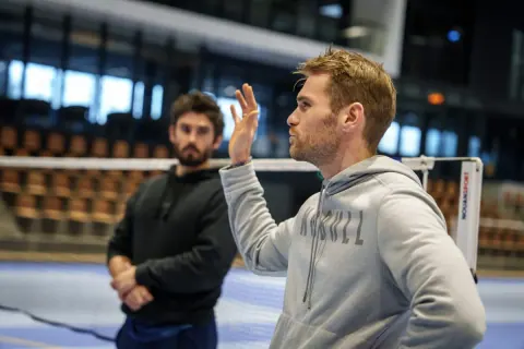Laurent Constantin, la main levée, explique un point de jeu à un joueur qui l’écoute au bord du filet, tribunes en arrière-plan